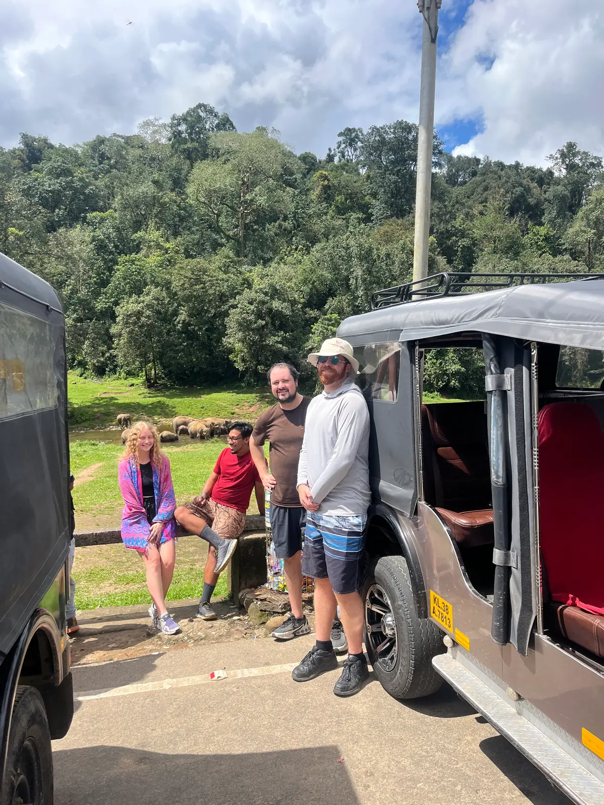 Travellers standing beside a Munnar Trip Makers jeep while wild elephants graze by the river